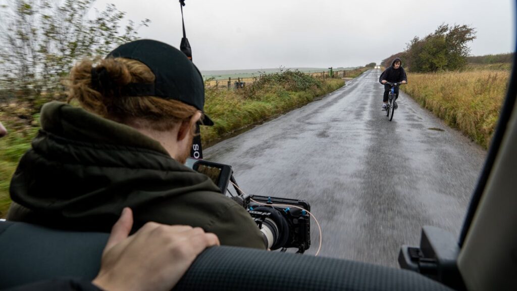 A man shooting a shot of a man on a bike on a rainy street.